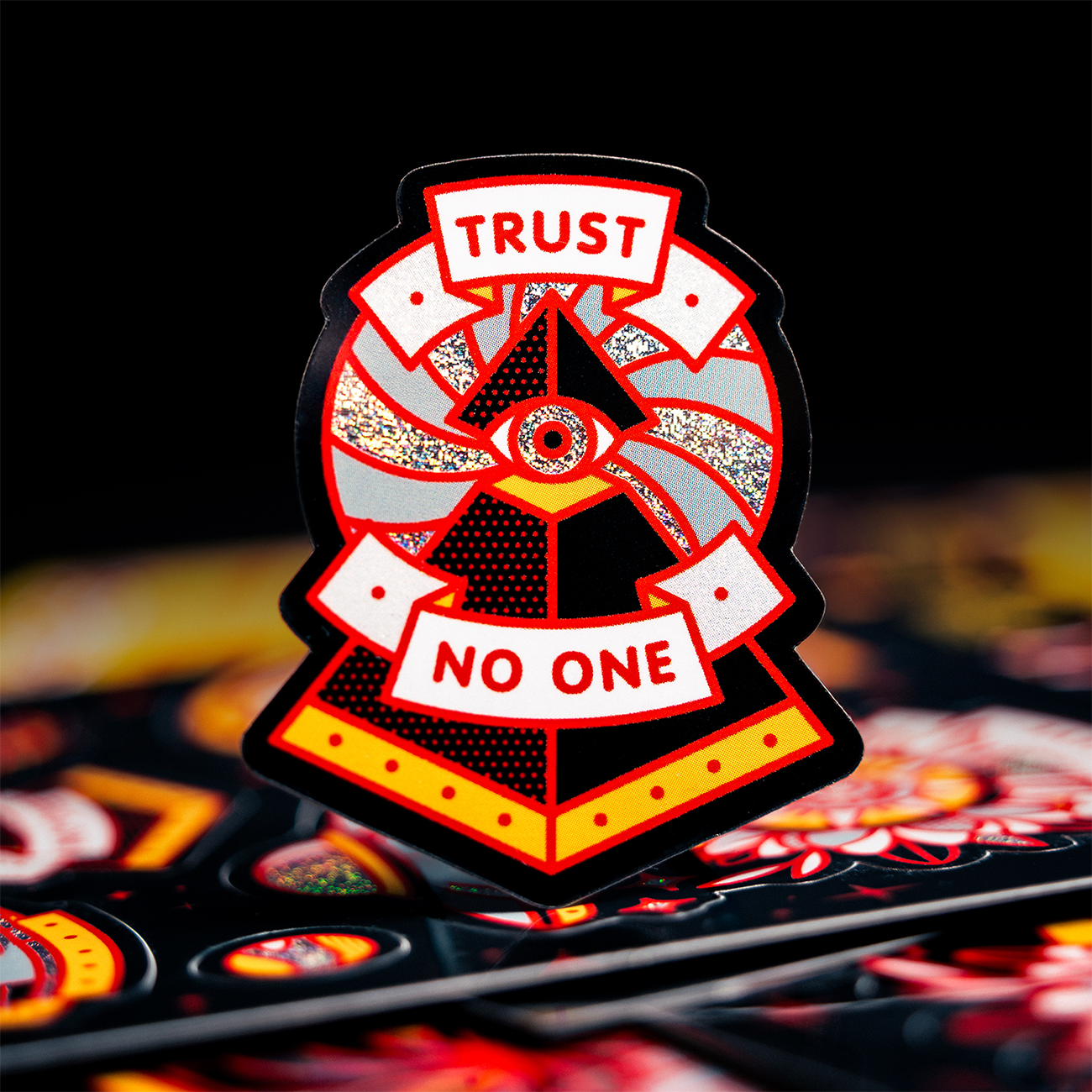 A pixie dust die-cut sticker with a red and black design featuring an eye and the text "Trust No One".