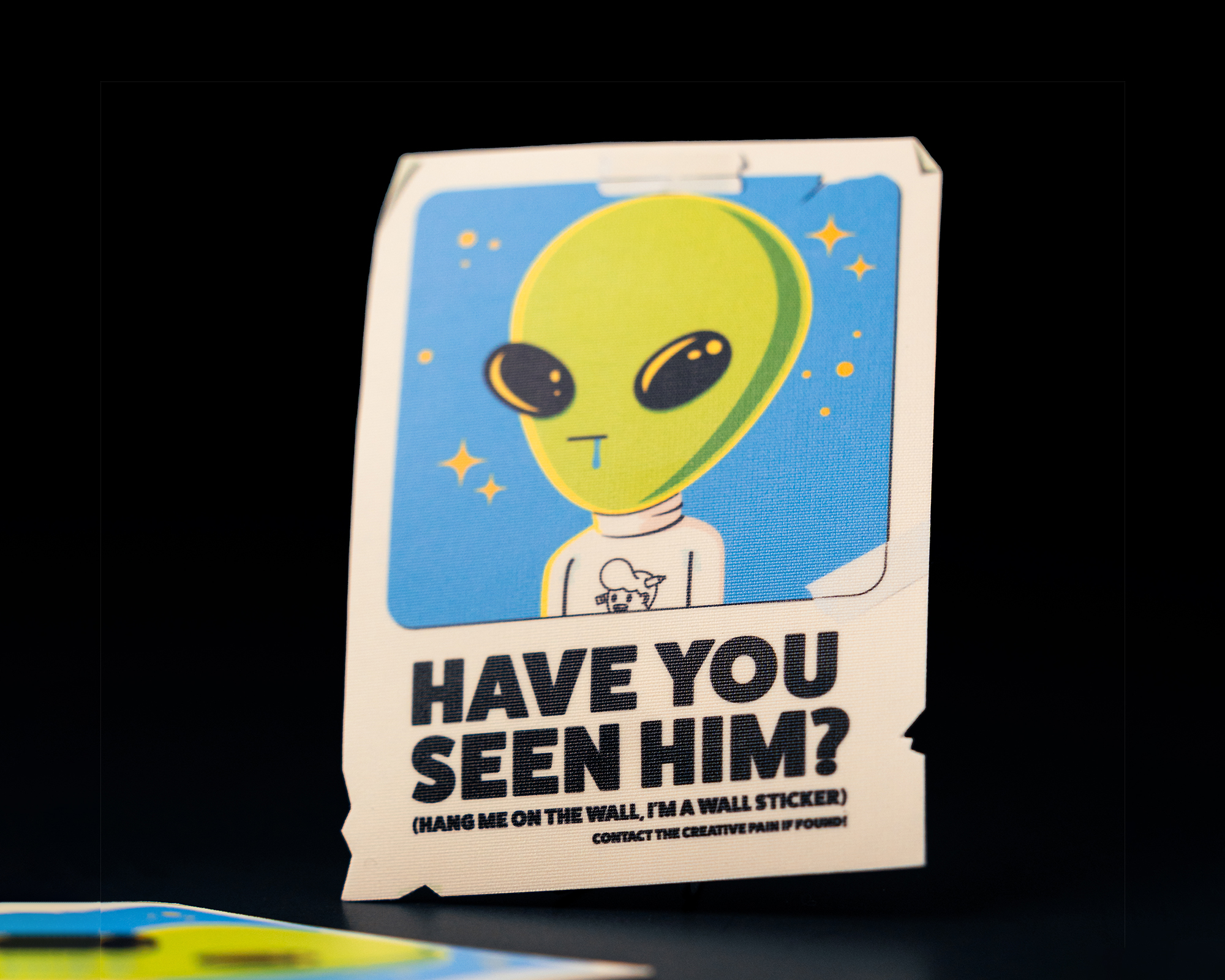 A wall sticker of a green alien with "Have You Seen Him?" text, set against a blue background, with instructions to hang it on a wall.