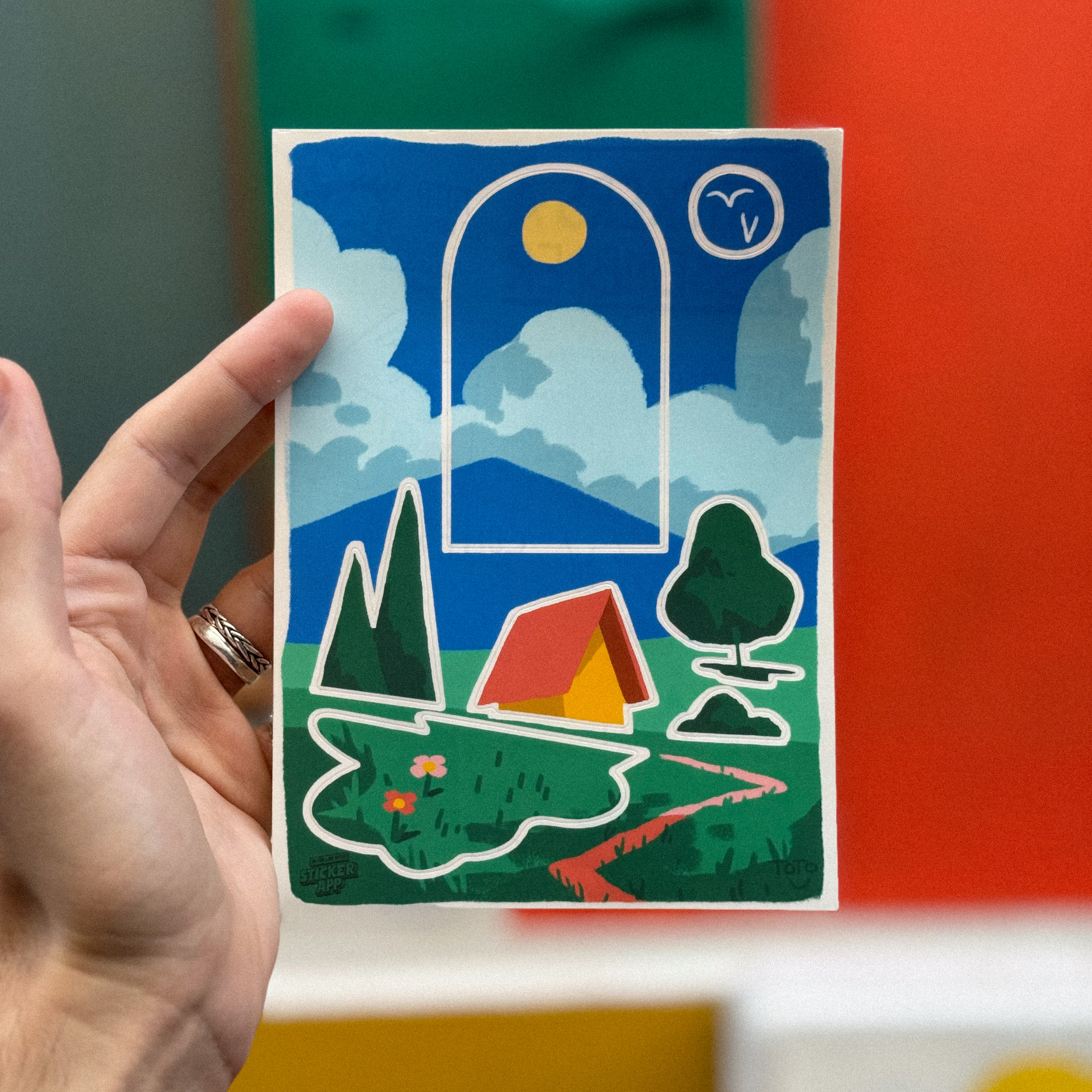 Hand holding a colorful sticker sheet of a landscape with a house, trees, mountains, and a large window framing a sun and clouds.
